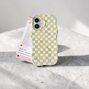 May include: A yellow and white checkered phone case with a wavy edge. A pink social media-style card is propped up behind the phone case. The card has a heart icon, a comment icon, and a profile icon.