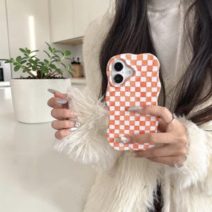 May include: An orange and white checkered phone case. The case has a wavy, irregular border. The phone case is held in front of a white background. The case has two camera lenses.