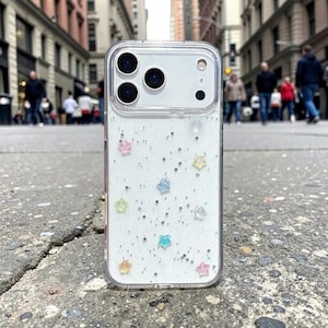 May include: Clear phone case with a white glitter background and colorful star accents. The case has a clear border and is designed to fit a smartphone. The case is shown outdoors on a gray surface.