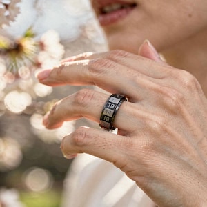 May include: A black ring with white numbers engraved on the band. The ring is worn on a finger. The numbers are 6, 7, 14, and 19. The ring is made of metal and has a shiny finish. The background is blurred.