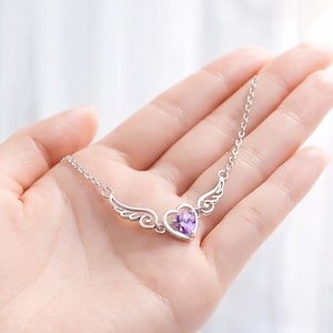 May include: A silver necklace featuring a heart-shaped pendant with a purple gemstone center, framed by delicate wing designs. The necklace is held in a hand, showcasing its intricate details and elegant design. The chain is a delicate silver.