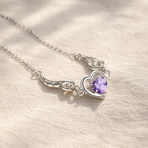 May include: A silver necklace featuring a heart-shaped pendant with a vibrant purple gemstone. The pendant is framed by a delicate silver design with wing-like accents. The chain is a fine, linked style.