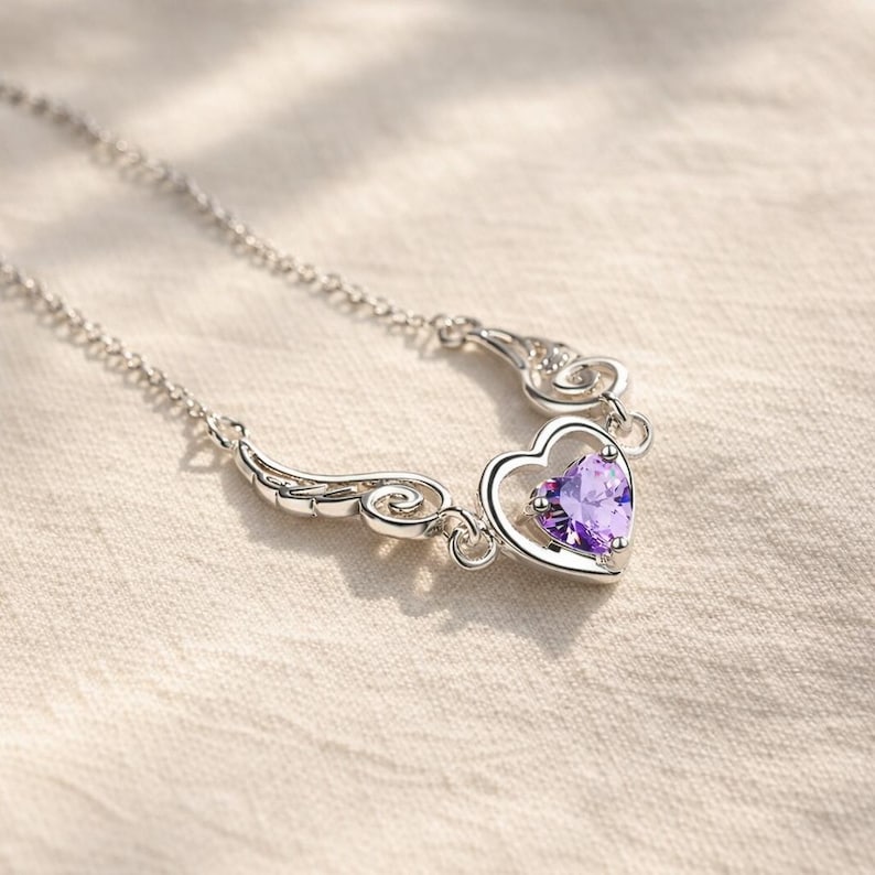 May include: A silver necklace featuring a heart-shaped pendant with a vibrant purple gemstone. The pendant is framed by a delicate silver design with wing-like accents. The chain is a fine, linked style.