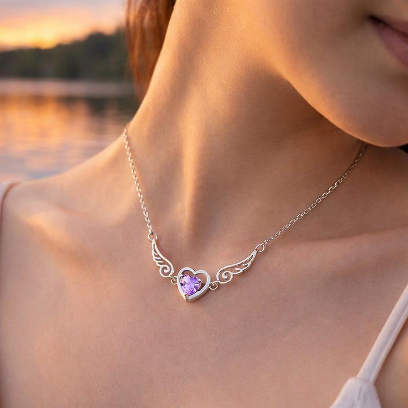 May include: A silver necklace featuring a heart-shaped pendant with a purple gemstone. The pendant is flanked by stylized wing designs. The necklace is worn around the neck.