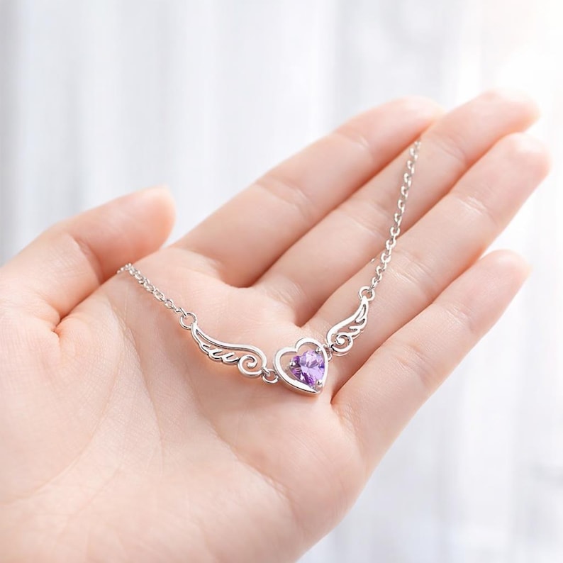 May include: A silver necklace featuring a heart-shaped pendant with a purple gemstone center, framed by delicate wing designs. The necklace is held in a hand, showcasing its intricate details and elegant design. The chain is a delicate silver.