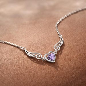 May include: A silver necklace featuring a heart-shaped purple gemstone pendant, framed by delicate wing designs. The necklace has a fine chain and is set against a warm brown background, showcasing the jewelry's elegant details.