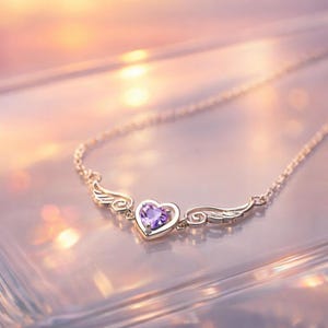 May include: A gold-colored necklace with a heart-shaped purple gemstone pendant. The pendant is framed by delicate wings. The necklace is displayed on a reflective surface with a soft, blurred background.