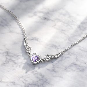 May include: A silver necklace featuring a heart-shaped pendant with a purple gemstone. The pendant is flanked by stylized wing designs. The necklace is displayed on a marble surface.