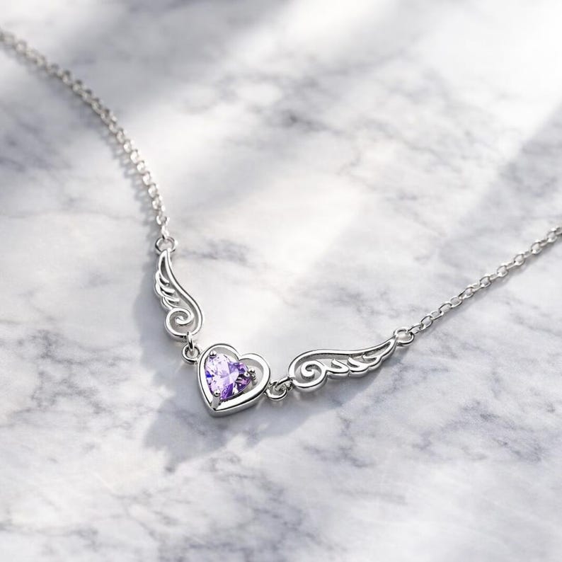 May include: A silver necklace featuring a heart-shaped pendant with a purple gemstone. The pendant is flanked by stylized wing designs. The necklace is displayed on a marble surface.