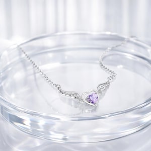 May include: A silver necklace with a heart-shaped pendant and wing accents. The heart features a purple gemstone. The necklace is displayed on a clear, reflective surface.