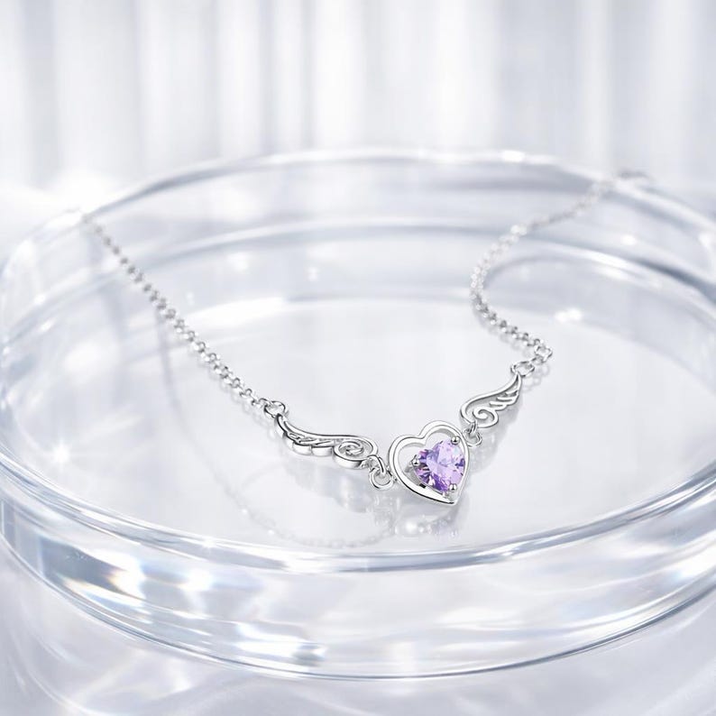 May include: A silver necklace with a heart-shaped pendant and wing accents. The heart features a purple gemstone. The necklace is displayed on a clear, reflective surface.