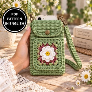 May include: A sage green crochet phone bag featuring a central granny square motif with a white and yellow flower. The bag has a flap with a bronze-coloured snap closure, a matching crochet strap, and a black circular label stating PDF PATTERN IN ENGLISH.