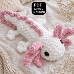 Może przedstawiać: Ręcznie robiona biało-różowa pluszowa zabawka axolotl. Axolotl ma różowe skrzela, różową płetwę grzbietową i czarne oczy. Tekst "PDF PATTERN IN ENGLISH" znajduje się w czarnym polu. Zabawka leży na kremowym kocu.