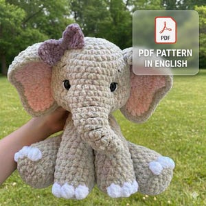 Può includere: Un peluche a forma di elefante all'uncinetto in tonalità di grigio, con orecchie interne rosa e un fiocco viola. L'elefante ha dettagli bianchi sui piedi e un'espressione amichevole. L'immagine include il testo "PDF PATTERN IN ENGLISH".