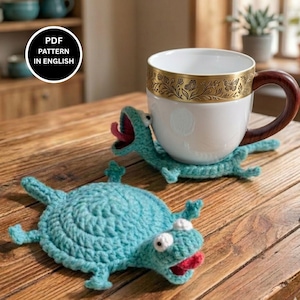 Puede incluir: Dos posavasos de ganchillo azul claro con forma de tortuga, ojos blancos y lenguas rojas. Uno sostiene una taza de cerámica blanca con una banda dorada de mariposas y asa marrón. Una insignia negra circular indica PDF PATTERN IN ENGLISH.