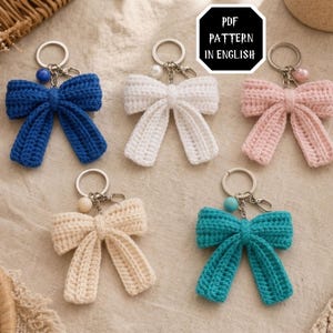 May include: Five crochet bow keychains in blue, white, light pink, beige, and teal. Each bow features a metal keyring with a small colored bead attached. A black sign with white text reads PDF PATTERN IN ENGLISH, indicating a digital file.