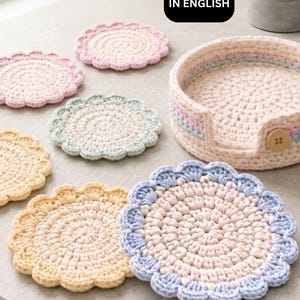 May include: A collection of handmade crochet items in pastel colors. Includes round coasters with scalloped edges in yellow, pink, blue, and green, and a matching basket. The text "PDF PATTERN IN ENGLISH" is in a black box.