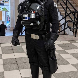 Peut inclure: Un costume de pilote de chasseur TIE noir, avec casque, boîtier pectoral, gants et bottes. Le costume comprend une combinaison noire et une ceinture. Le casque a un design distinctif avec un emblème central.