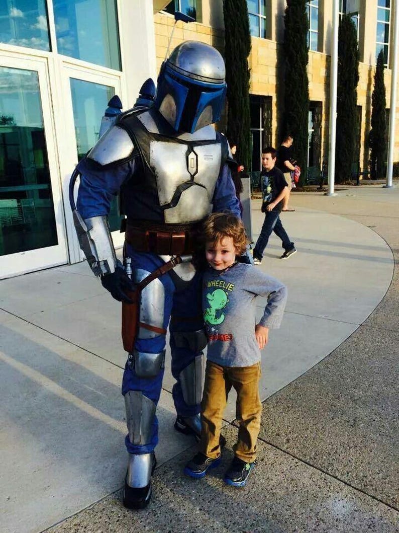 K&ouml;nnte beinhalten: Eine Person in einem silber-blauen Ganzk&ouml;rper-R&uuml;stungskost&uuml;m im Mandalorianer-Stil mit Helm und Jetpack, die neben einem Kind steht, das ein graues Langarmshirt mit einem gr&uuml;nen Dinosaurier-Motiv und dem Text WHEELIE sowie eine braune Hose tr&auml;gt.