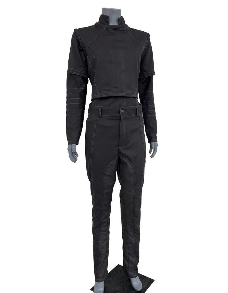 May include: A black two-piece outfit consisting of a long-sleeved cropped top with a high collar and matching trousers. The top features horizontal stitching on the sleeves, while the trousers have reinforced panels and visible seam detailing.