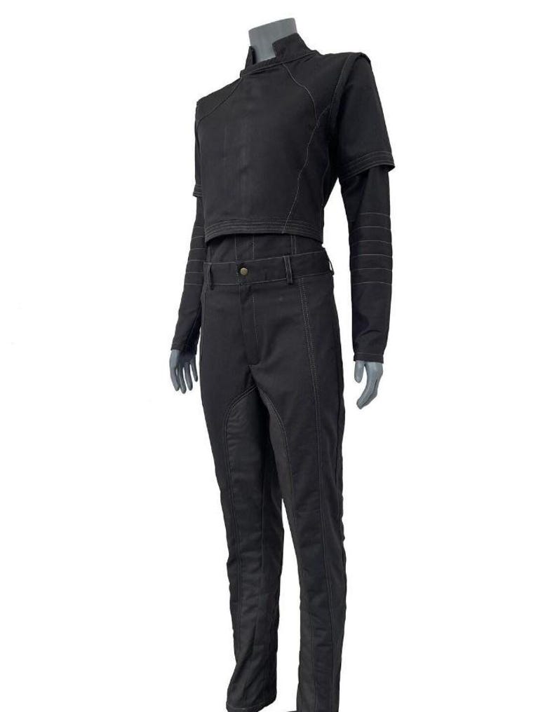 May include: A black two-piece outfit consisting of a cropped long-sleeve top and matching trousers. The top features a high collar and structured shoulders, while the pants have a fitted waistband and visible seam detailing throughout the garment.