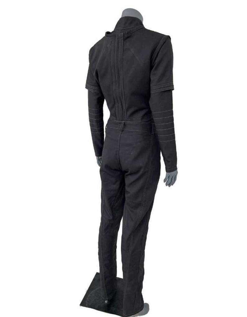 May include: A black long-sleeved jumpsuit featuring a high collar, structured shoulders, and visible white topstitching along the seams. The garment includes a fitted bodice, a defined waistline with belt loops, and straight-leg trousers.