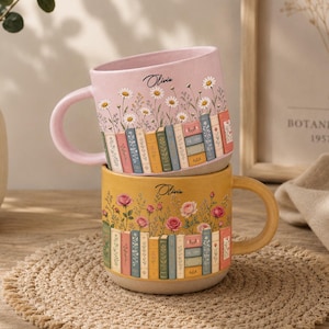 Peut inclure: Deux tasses en céramique empilées avec un motif imprimé de livres colorés et de fleurs. La tasse supérieure est rose clair avec des marguerites, et la tasse inférieure est jaune avec des roses. Le prénom Olivia est inscrit en noir au-dessus.