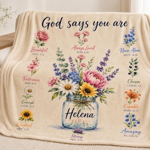 May include: A cream-coloured fleece blanket featuring a central glass mason jar filled with colourful flowers. The text reads God says you are, Helena, and various inspirational words with Bible verses paired with floral illustrations around the jar.