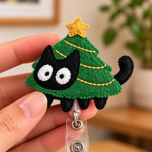 May include: A felt badge reel featuring a black cat shaped like a green Christmas tree with a yellow star on top. The cat has large white eyes with black pupils, and the tree is decorated with gold stitched lines. The reel has a clear plastic strap.