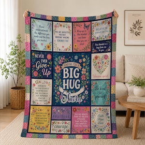 Può includere: Una coperta rettangolare con bordo patchwork e diversi pannelli con motivi floreali e citazioni ispiratrici. Il pannello centrale mostra il testo 'SENDING YOU A BIG HUG' con il nome 'Olivia' e un'illustrazione di braccia.