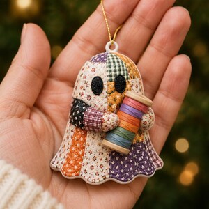 May include: A ghost-shaped ornament featuring a patchwork quilt pattern with various floral and plaid fabrics in orange, purple, and white. The ghost has two black oval eyes and holds a wooden spool of thread wrapped in rainbow-colored yarn.