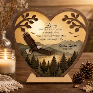 May include: A heart-shaped wooden memorial plaque featuring a laser-cut eagle in flight over a forest and lake scene. The text reads: Love NEVER TRULY LEAVES it simply rises. NOW YOU SOAR WHERE ONLY angels and eagles fly. Olivia Smith 1977-2025.