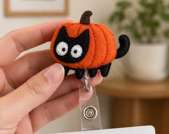 Pode incluir: Um porta-crachá de feltro com um rosto de gato preto com grandes olhos brancos, vestindo um traje de abóbora laranja com um caule castanho e uma cauda preta. A figura do gato está presa a um cordão retrátil de plástico transparente.