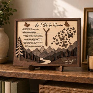 May include: A rectangular wooden memorial plaque featuring a layered mountain landscape, a pine tree, and butterflies. The text reads As I Sit In Heaven and In Loving Memory of Daniel Taylor 1960 2024. It includes a central Y-shaped branch.