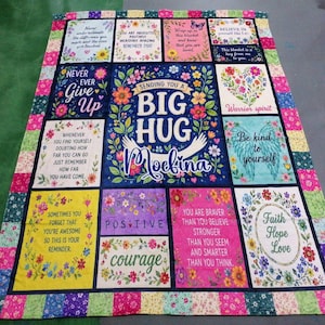 May include: A rectangular quilt featuring a central panel with the text "SENDING YOU A BIG HUG Moelina" surrounded by floral motifs. The quilt is composed of multiple square and rectangular blocks with various inspirational quotes and colorful patterns.