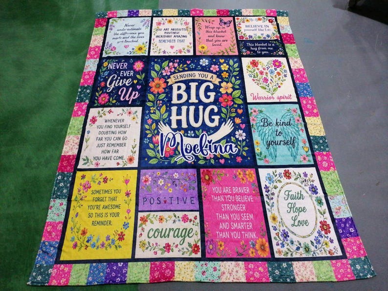 May include: A rectangular quilt featuring a central panel with the text "SENDING YOU A BIG HUG Moelina" surrounded by floral motifs. The quilt is composed of multiple square and rectangular blocks with various inspirational quotes and colorful patterns.