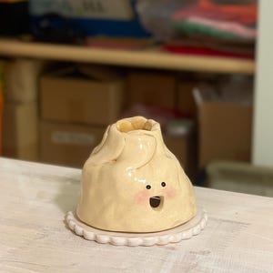 May include: A quirky, handmade ceramic dumpling-shaped container with a smiling face and pink cheeks. The container sits on a white, scalloped-edge plate. The dumpling is a light beige colour.