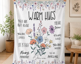 May include: A white fleece blanket featuring a central bouquet of colorful wildflowers and the text "WARM HUGS" at the top. The blanket includes inspirational phrases like "YOU ARE NOT ALONE," "YOU ARE BRAVE," and the name "Olivia" in script.