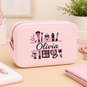 May include: A rectangular pink and white striped cosmetic bag featuring an embroidered design of makeup brushes, a perfume bottle, and compacts. The name Olivia is embroidered in black script across the center of the design.