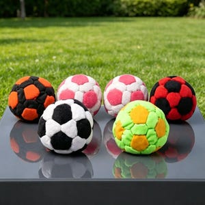 May include: A collection of six colorful soccer balls. The balls feature various color combinations, including black and white, red and black, orange and black, pink and white, and green and orange. They are arranged on a reflective surface with a grassy background.