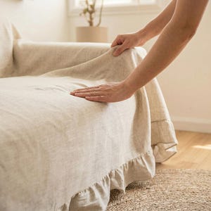 Puede incluir: Funda de sofá de lino beige con falda con volantes. La mano de una persona alisa la funda. El sofá está en una habitación con una paleta de colores neutros y luz natural.