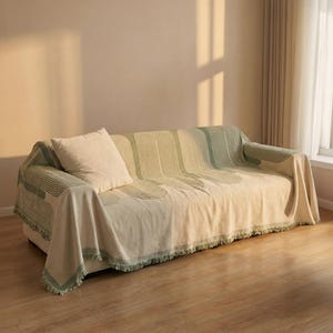 Op de afbeelding: Een beige bankdeken met een groen geometrisch patroon en franjes. Een bijpassend beige kussen ligt op de bank. De deken heeft een modern, minimalistisch ontwerp.