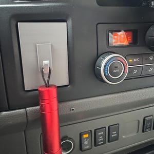 May include: A red cylindrical object hangs from a silver rectangular plate attached to a vehicle's dashboard. The dashboard features climate controls, including a digital display showing 98°F, and various buttons.