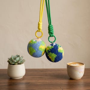 Puede incluir: Dos adornos de globo de ganchillo, uno con un cordón y un anillo amarillos, el otro con verde. Los globos presentan hilos azules, verdes y amarillos, que representan continentes y océanos. Una suculenta en maceta y una taza de café están sobre una superficie de madera.