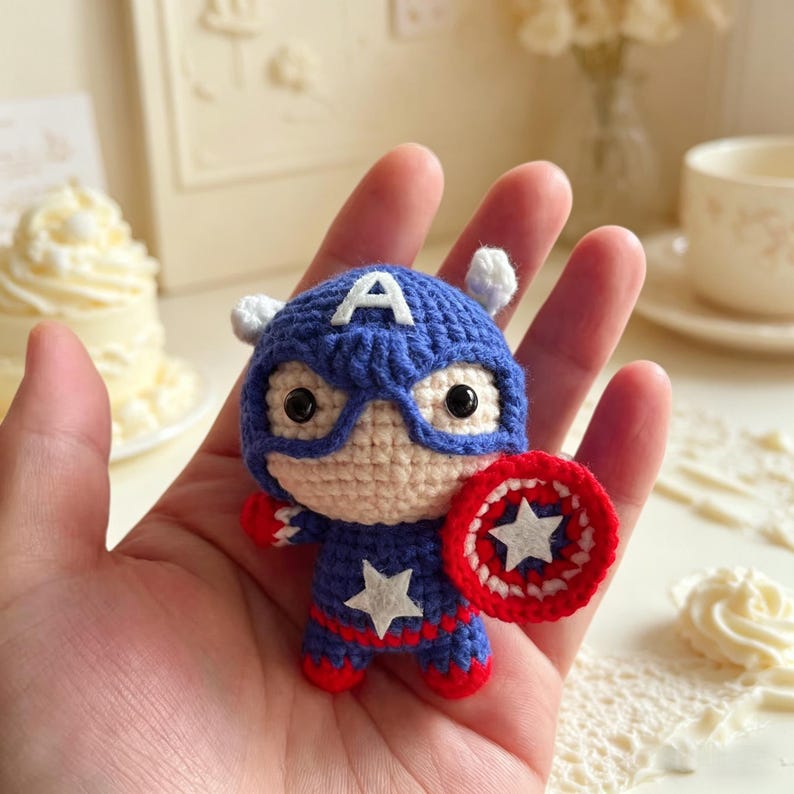 Hand holding a small handmade crochet star shield hero amigurumi doll with big eyes and blue suit in a cozy cream colored room