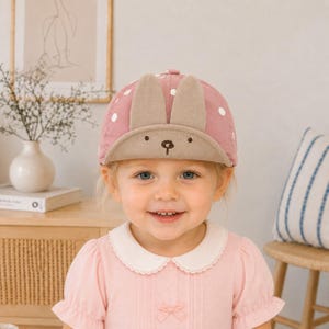Puede incluir: Una gorra de béisbol rosa empolvado con lunares blancos y dos orejas de conejo de tela beige erguidas en la parte delantera. La visera beige muestra una nariz y boca bordadas en marrón, creando un diseño sencillo de cara de animal.