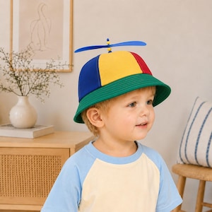 Puede incluir: Un sombrero de pescador multicolor con paneles de tela azul, amarilla y roja y un ala ancha de color verde. Una pequeña hélice de plástico azul con una cuenta central amarilla está fijada en la parte superior del sombrero, creando un accesorio lúdico.