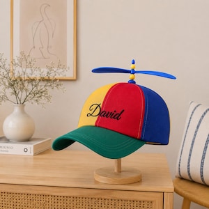 Puede incluir: Una gorra de béisbol multicolor con una corona de paneles amarillos, rojos y azules y una visera verde. El nombre David está bordado en cursiva negra en el panel rojo. Un juguete de hélice de plástico azul está fijado en la parte superior.