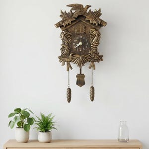 May include: A detailed, antique-style cuckoo clock in a warm brown finish, mounted on a white wall. The clock features intricate carvings, a bird on top, and decorative hanging weights. A wooden shelf below holds potted plants and a glass bottle.