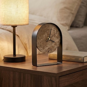 Peut inclure: Une horloge décorative moderne avec un cadran rond et texturé et des aiguilles noires. L'horloge est encadrée dans un cadre en métal noir. Une petite lampe avec un abat-jour beige est à gauche. L'horloge est sur une table de chevet en bois.
